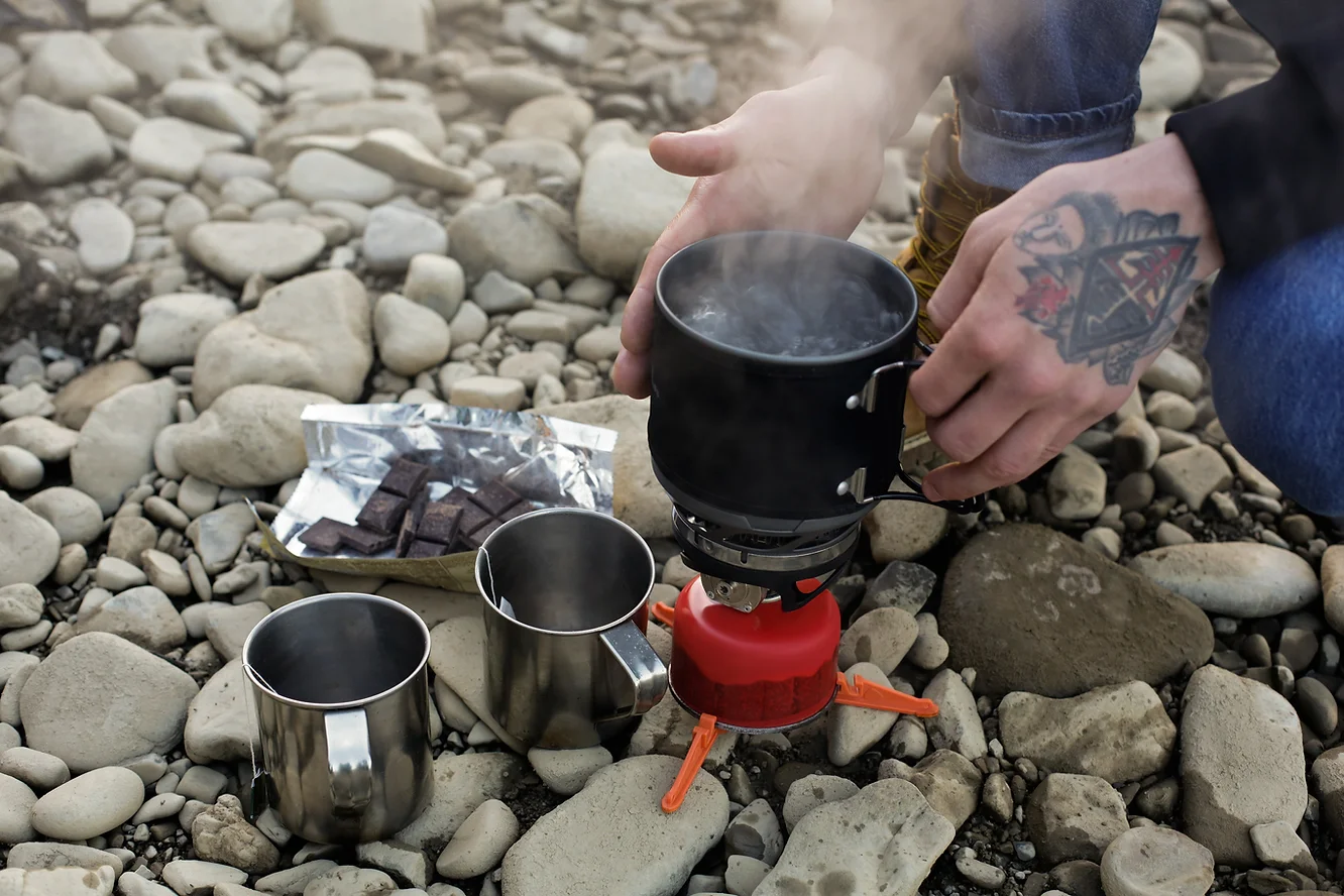 a person with a hand tattoo boils a pot of water using a portable camp stove.