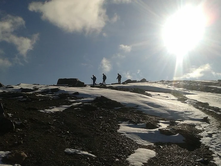a bright sun-saturated photo of a snow capped ridge, with the silhouettes of three hikers on top